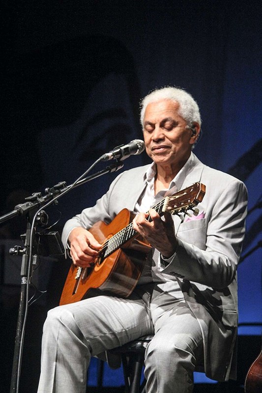 Paulinho da Viola de terno cinza, sentado, segurando um violão e cantando - Metrópoles