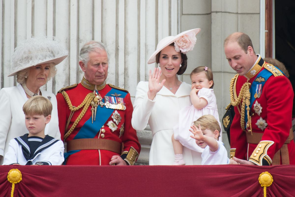 Foto colorida. Camilla Parker, rei Charles, Kate Middleton, princesa Charlotte, príncipe George e príncipe William. Eles estão em uma varanda. Os homens vestem trajes militares - família real britânica