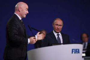 Em primeiro plano está Gianni Infantino, presidente FIFA, que discursa num pulpito, e Vladimir Putin, presidente da Rússia o escutando ao seu lado - Metrópoles
