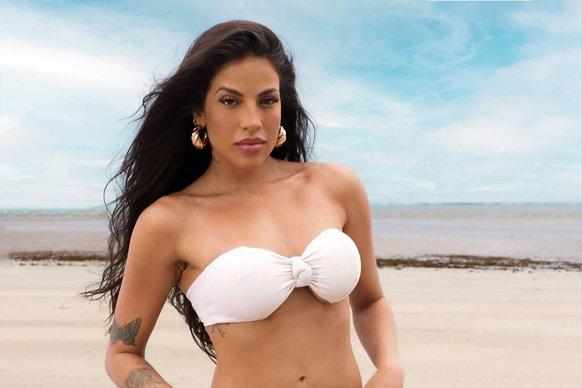 Na imagem colorida, mulher usa biquíni branco e posa na praia, com cabelos ao vento - Metrópoles