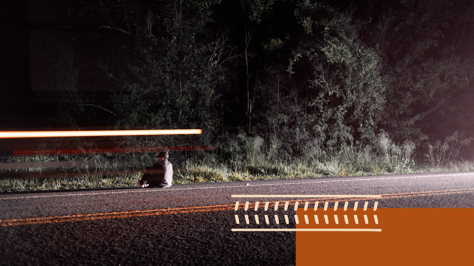 Foto mostra um jovem sentado à veira de uma estrada de costas