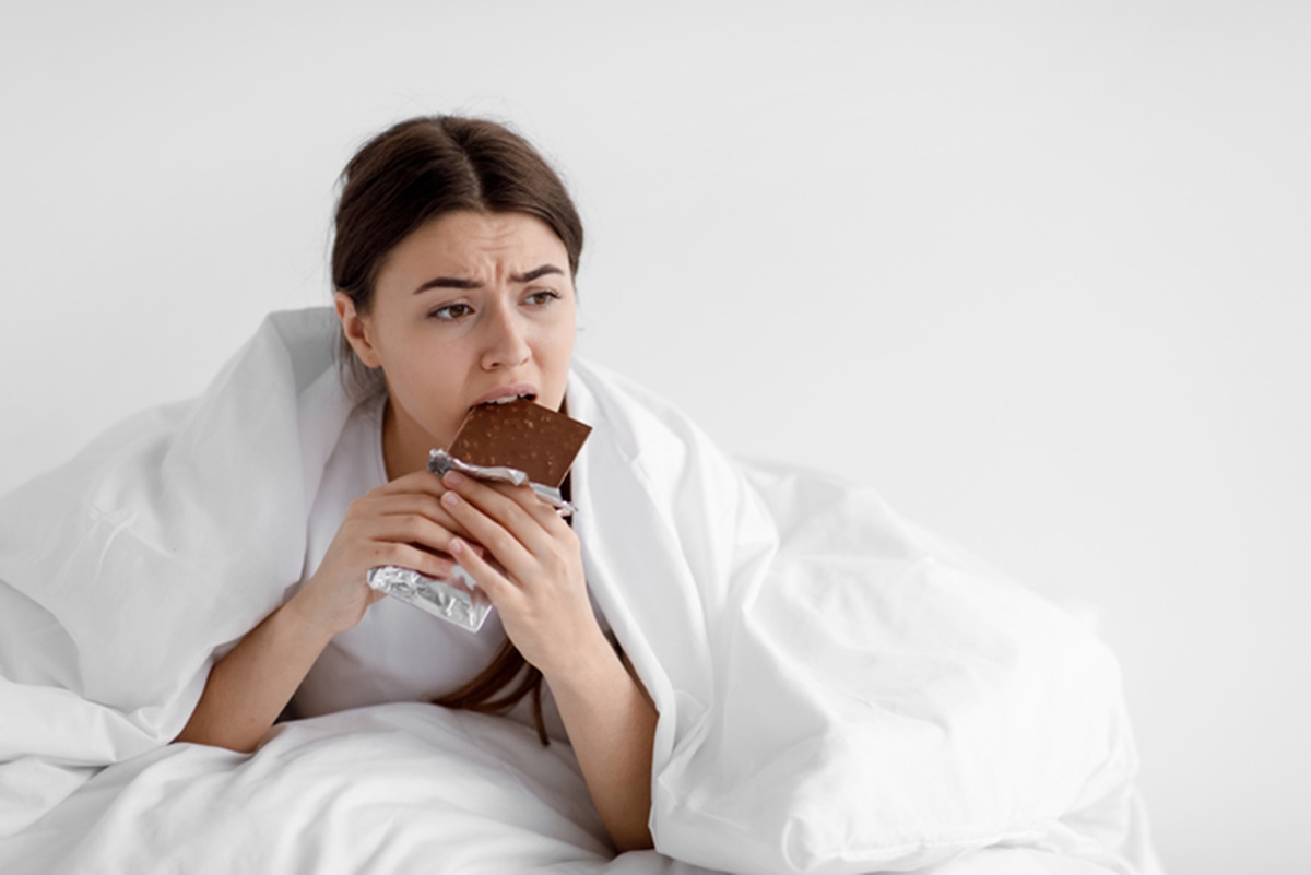 Foto colorida de mulher embaixo de cobertor comendo chocolate - Metrópoles