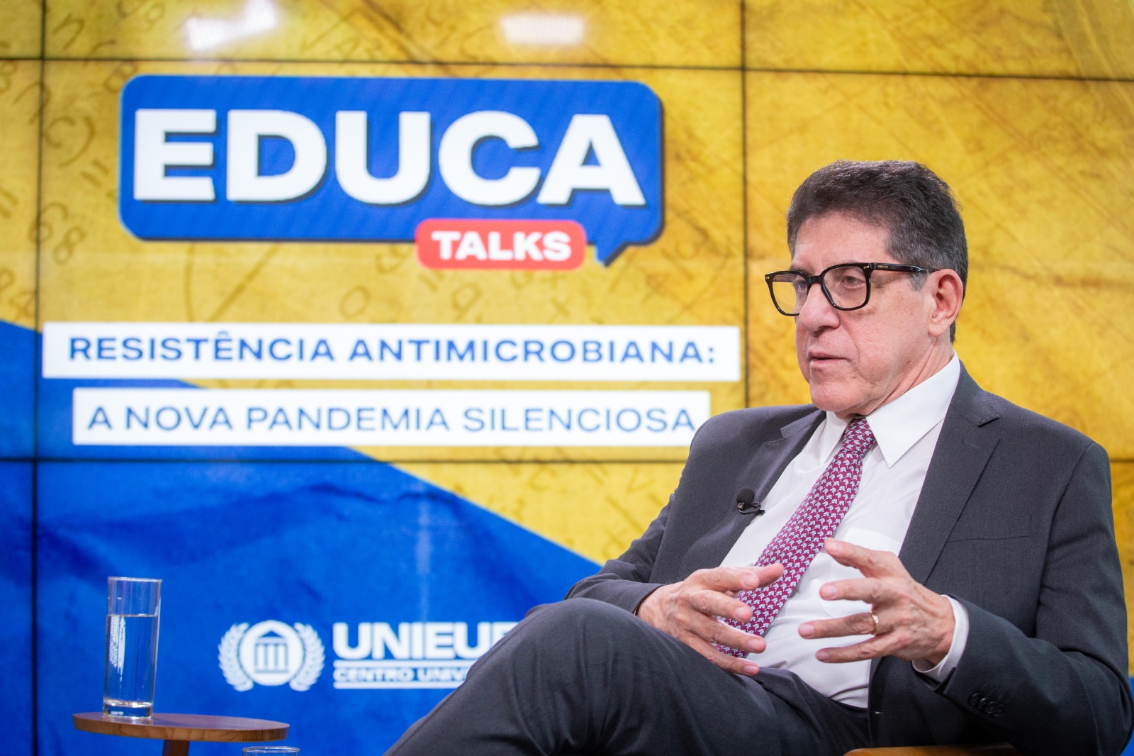 O infectologista, de terno escuro e óculos, está sentado e fala com as mãos entrelaçadas. Ao fundo, aparece o painel azul e amarelo com o logo do Educa Talks