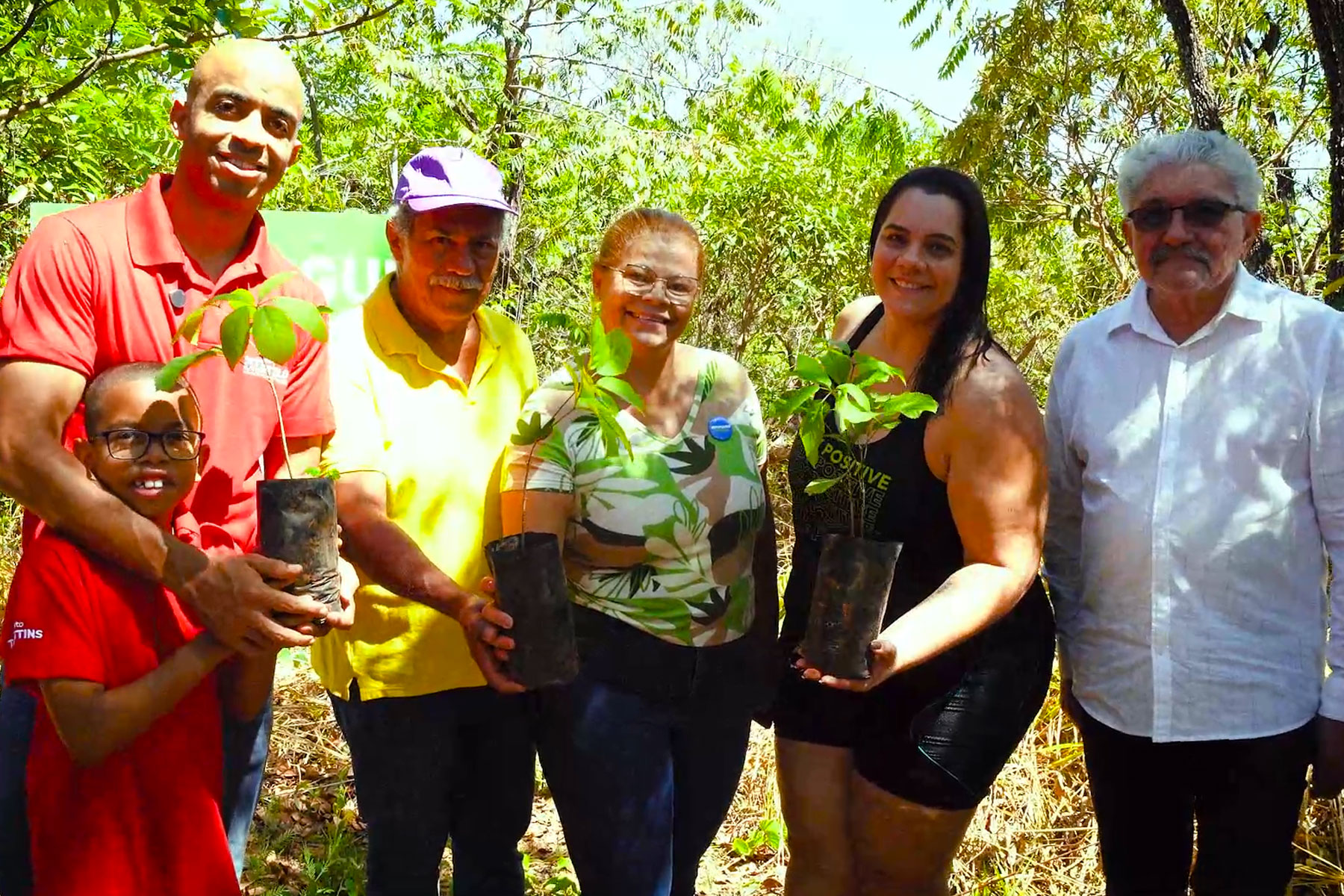 Cinco pessoas lado a lado, sorrindo e exibindo mudas de ipê, com árvores ao fundo