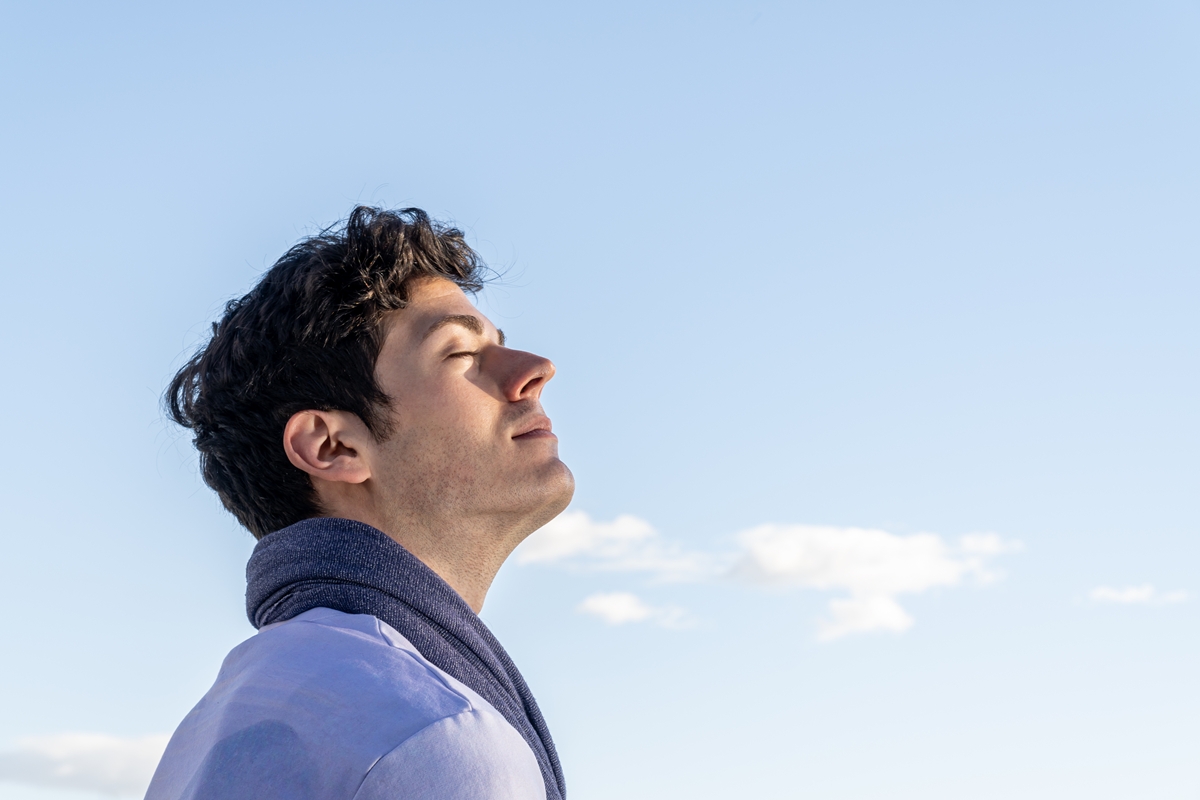 Retrato de um homem com a cabeça voltada para o céu e os olhos fechados em um dia claro, com céu azul e algumas nuvens brancas.