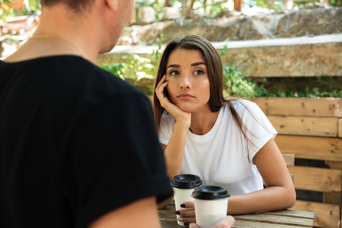 Jovem entediada sentada tomando café com o namorado em um café ao ar livre. Metrópoles