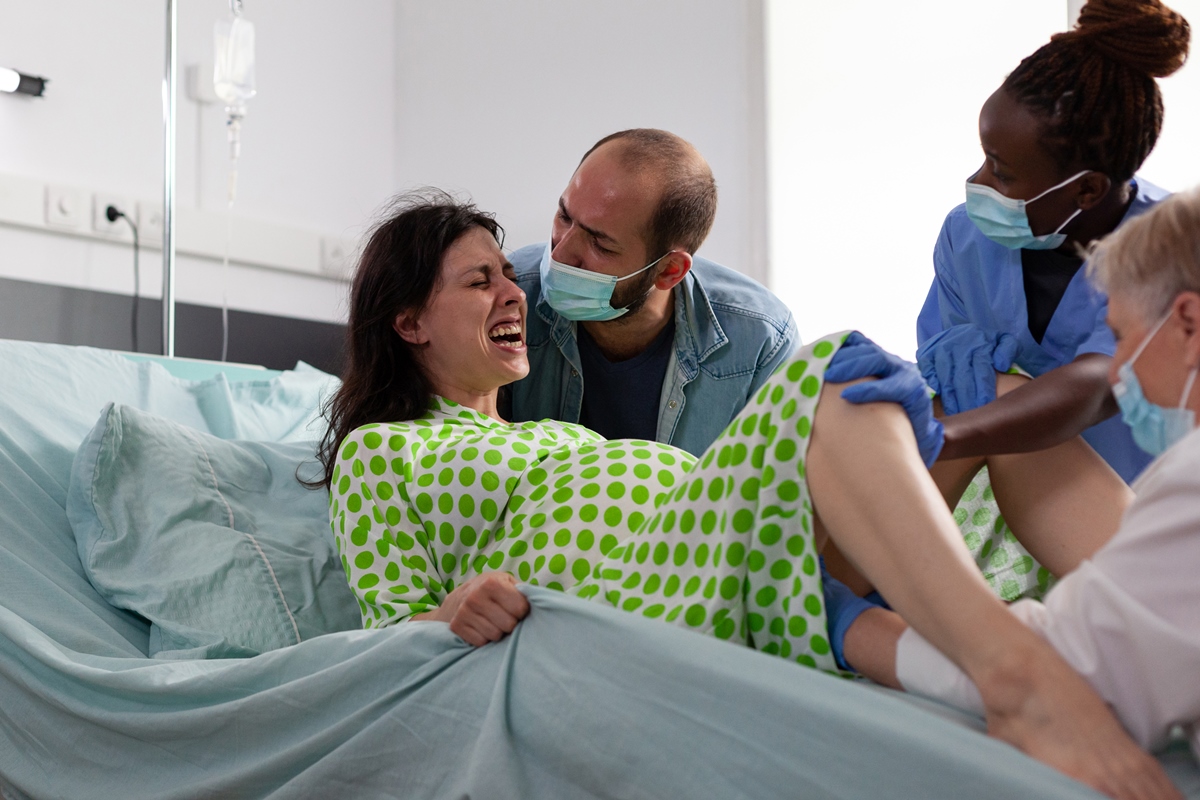 Foto colorida de mulher em trabalho de parto, deitada numa cama, acompanhada de médicos - Metrópoles