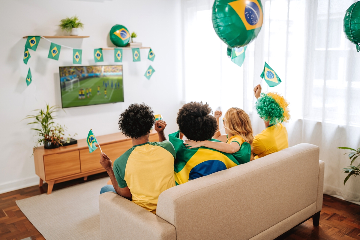 Foto colorida de pessoas sentadas em sofá assistindo ao jogo do Brasil. Tem decoração da Copa na sala de estar - Metrópoles