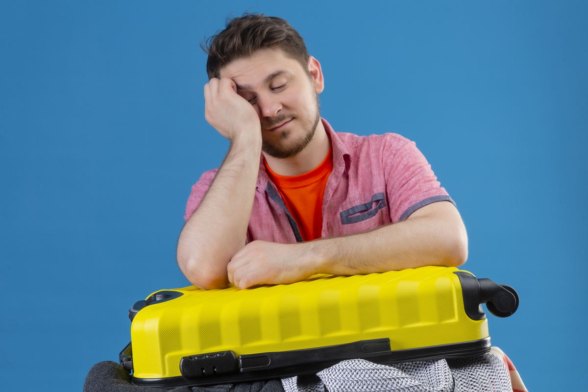 Foto colorida de homem cansado, com mala de viagem sobre o colo, mostrando cansaço nas férias - Metrópoles. 