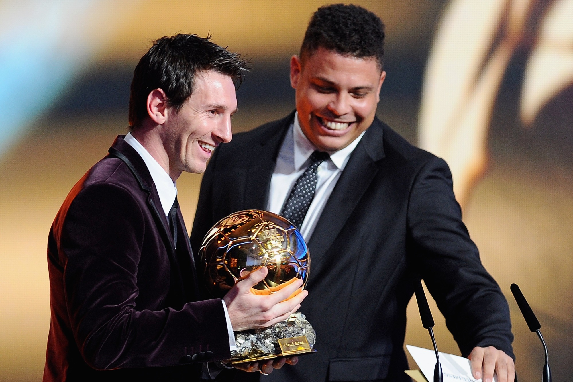 Foto colorida de dois homens de terno, um deles segurando a Bola de Ouro - Metrópoles