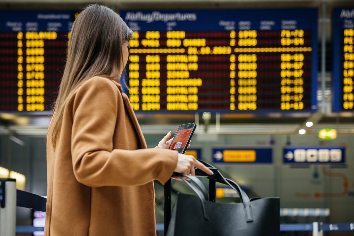 Foto colorida de mulher em aeroporto olhando o celular e segurando uma mala - Metrópoles