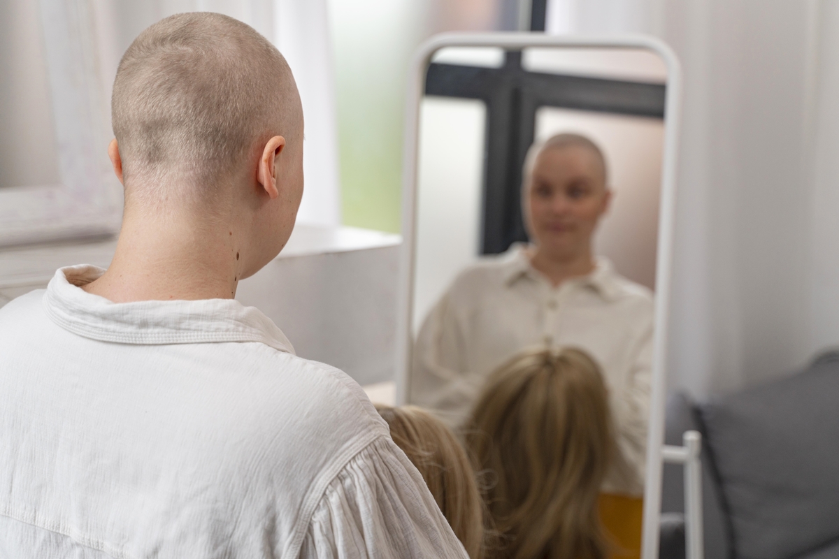 foto colorida sem foco de mulher careca em frente a um espelho segurando uma peruca, após tratamento de câncer - Metrópoles.