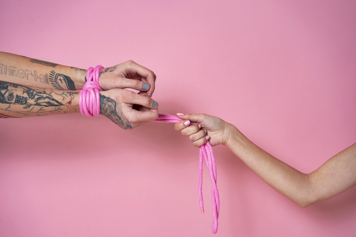 Foto colorida de mãos amarradas em fundo rosa