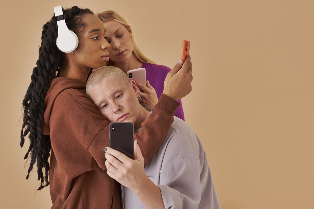 Foto colorida de meninas apoiadas umas nas outras enquanto estão com celular na mão