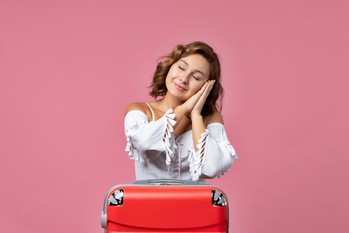 Foto colorida com mulher fazendo expressão de sono em cima de mala em fundo rosa