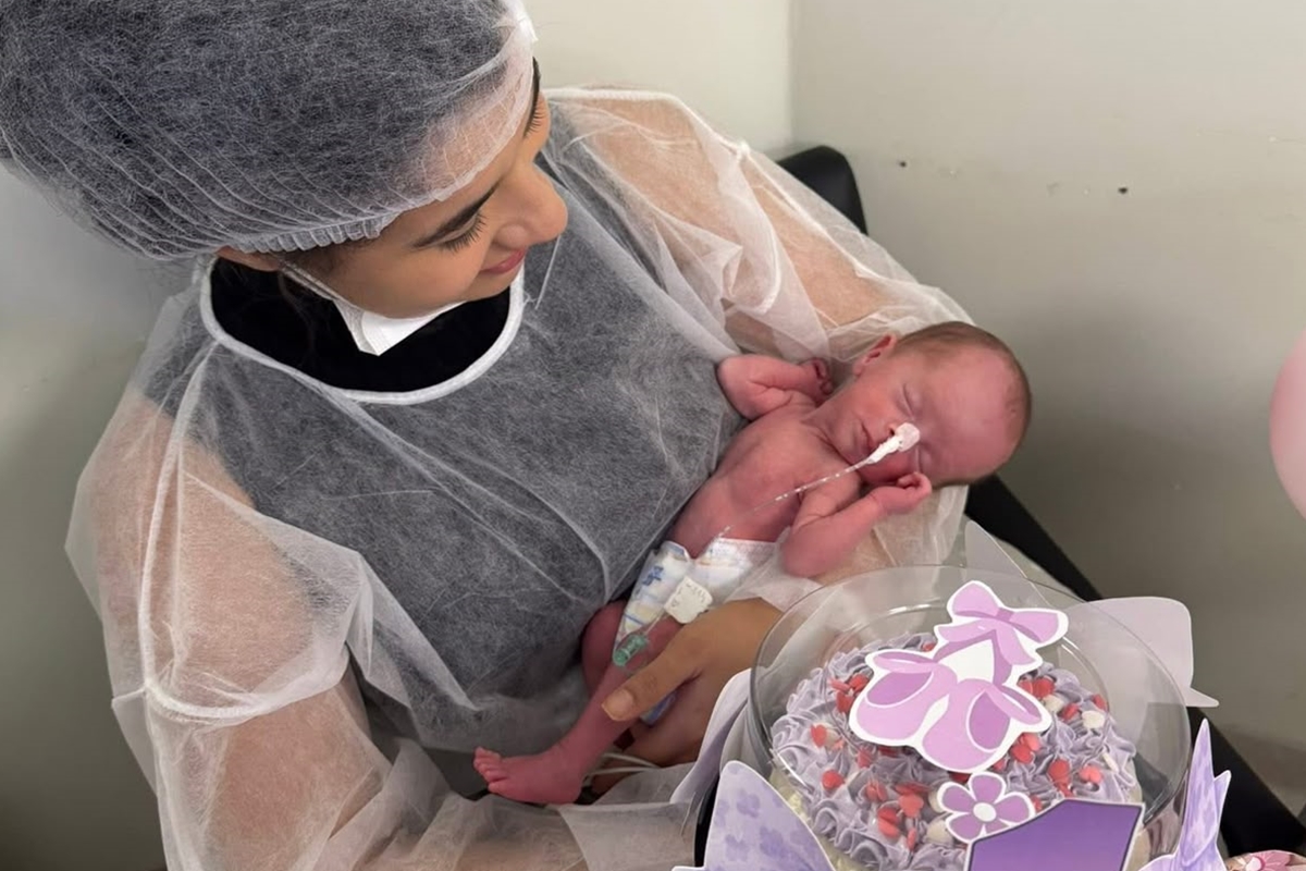 Foto colorida de mulher segurando bebê recém-nascida em ambiente hospitalar - Metrópoles.