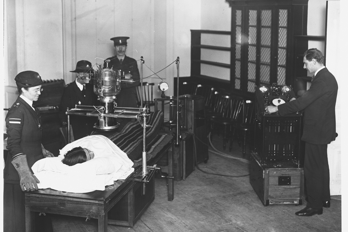 Imagem antiga mostra várias pessoas em pé enquanto paciente está deitado debaixo de um aparelho - Metrópoles