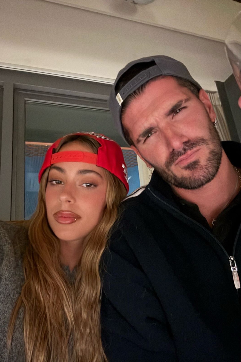 Foto colorida de mulher com boné vermelho e cabelo grande tirando foto com homem de boné preto - Metrópoles