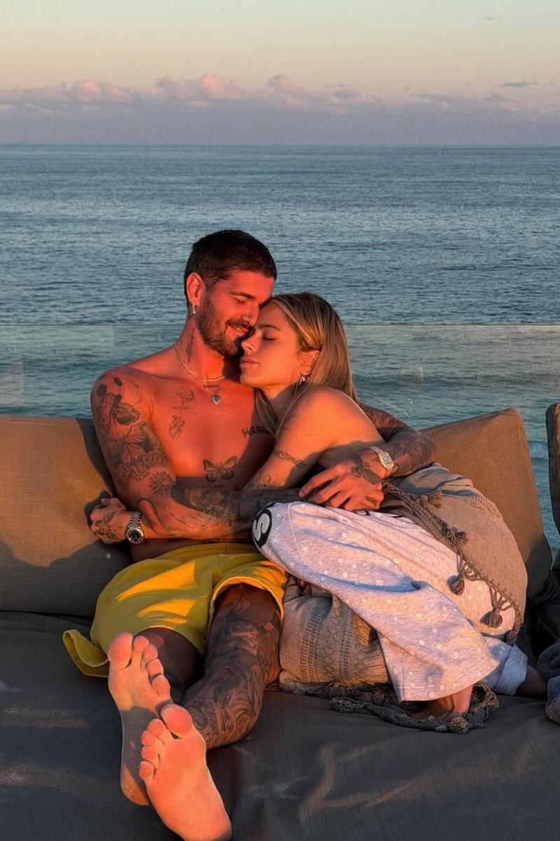 Foto colorida de mulher e homem deitados em uma cadeira de praia. Eles estão abraçados - Metrópoles