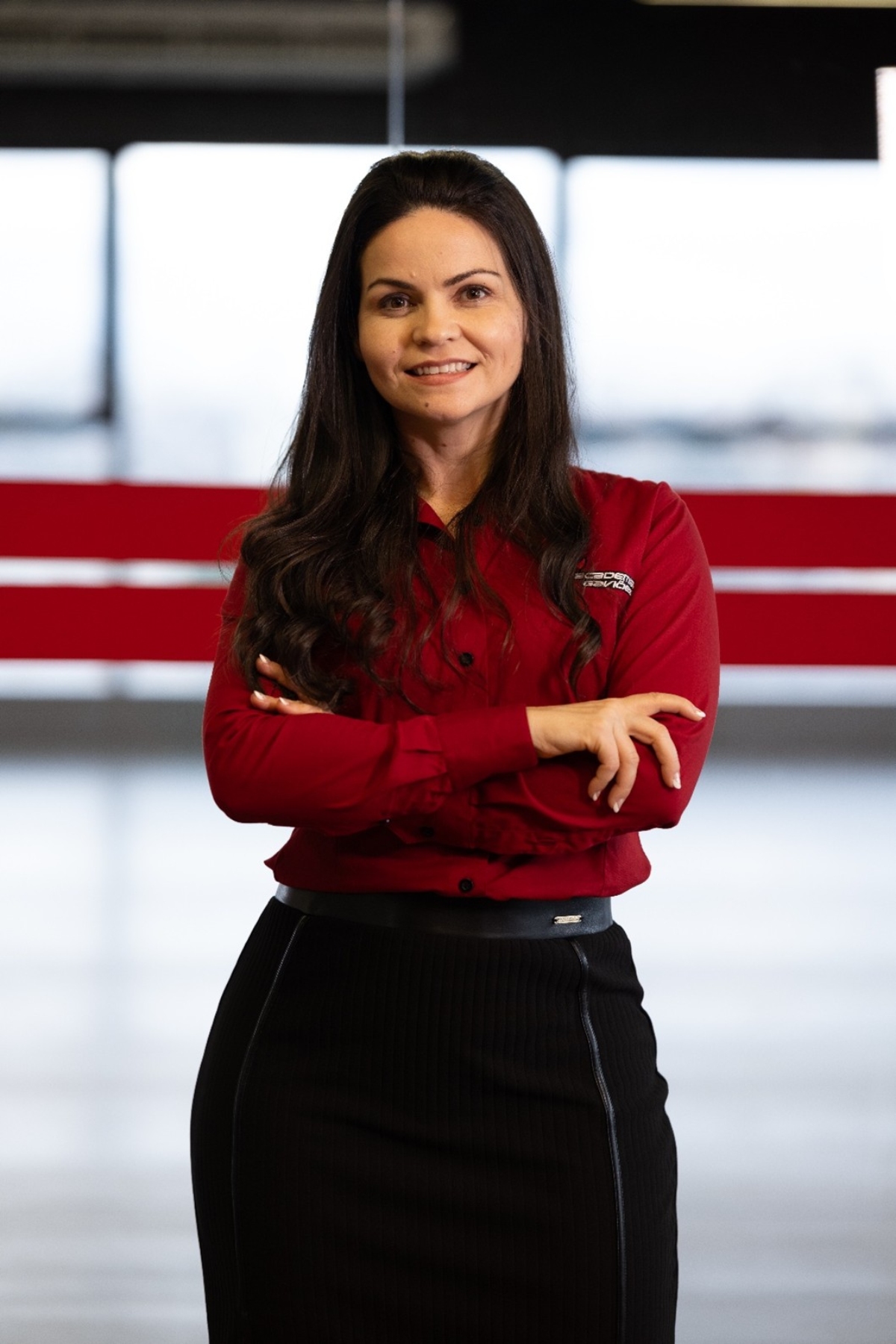 Retrato de Priscila Aguiar, CEO da Academia Gaviões 24h. Ela aparece em pé, sorrindo, com os braços cruzados, em um ambiente corporativo.
