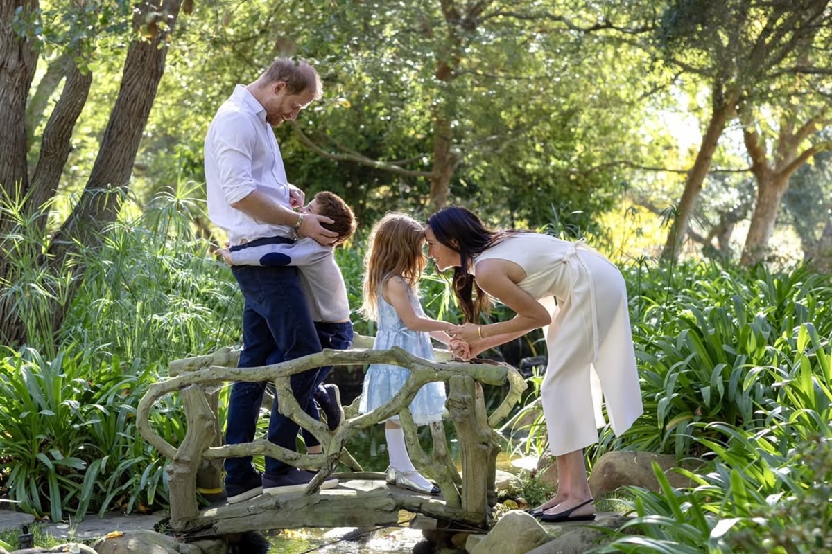 Foto colorida de homem e mulher com duas crianças em uma ponte situada em um jardim - Harry e Meghan - Metrópoles