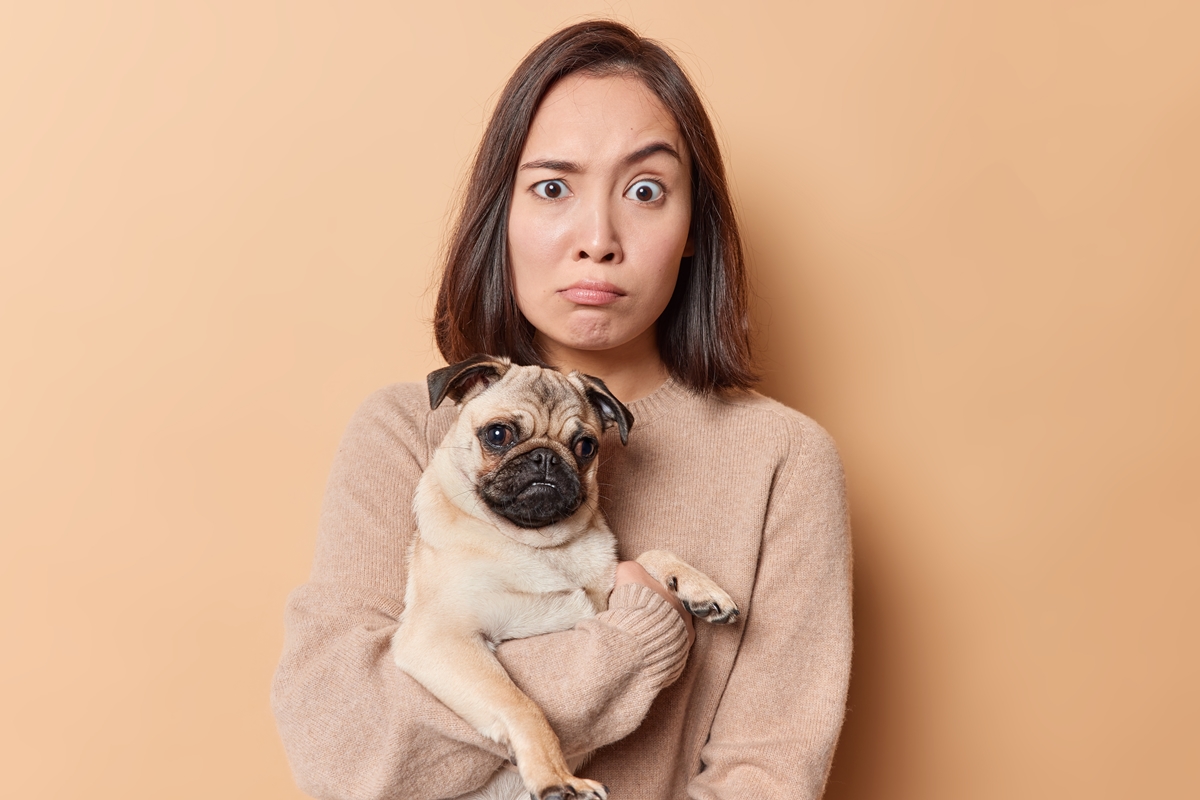 Foto colorida de mulher com expressão facial incomum, segurando um cachorro em seus braços - Metrópoles.