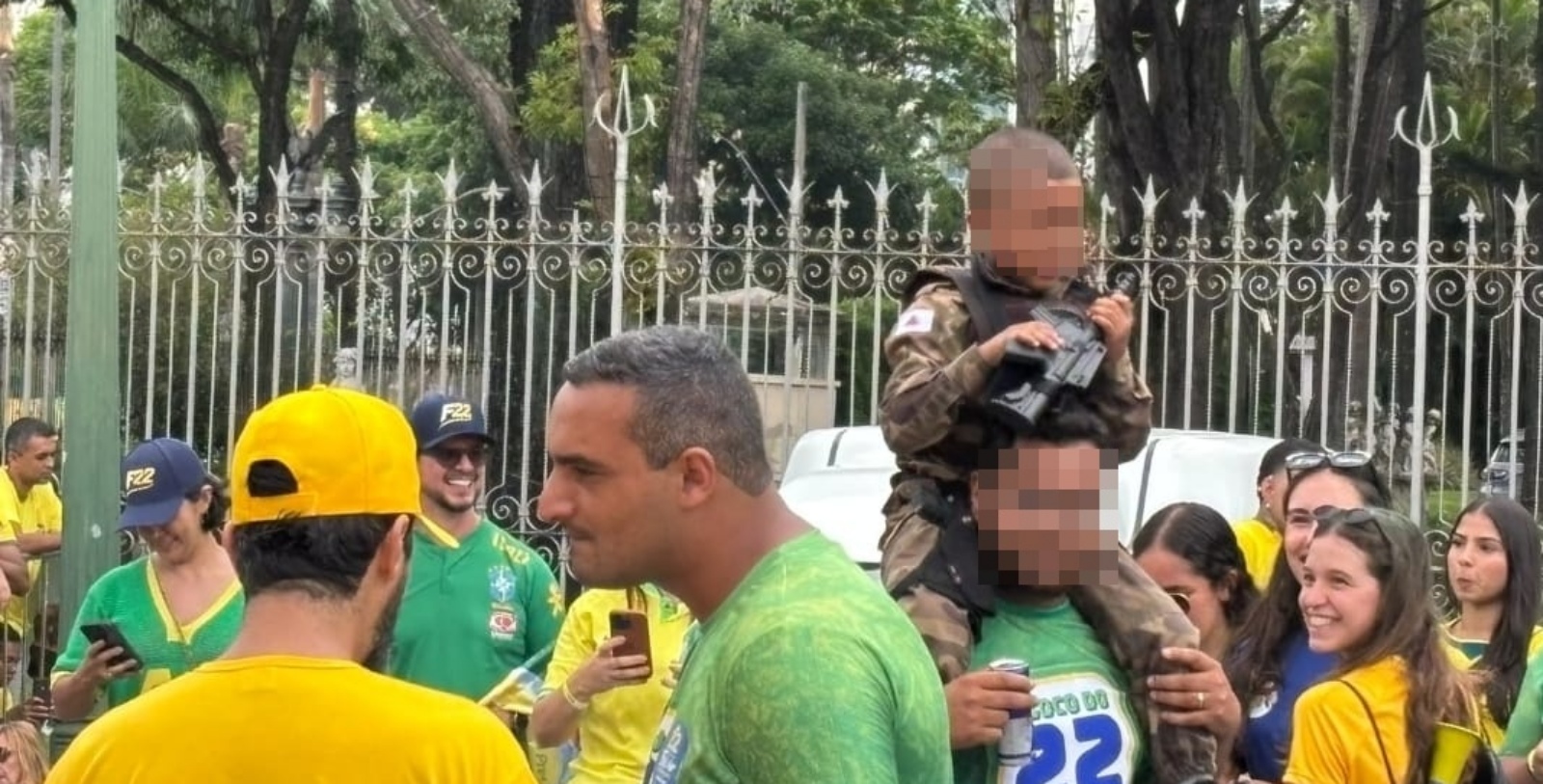 Criança de farda militar segura fuzil de brinquedo nos ombros de um homem em ato de Flávio Bolsonaro em Belo Horizonte