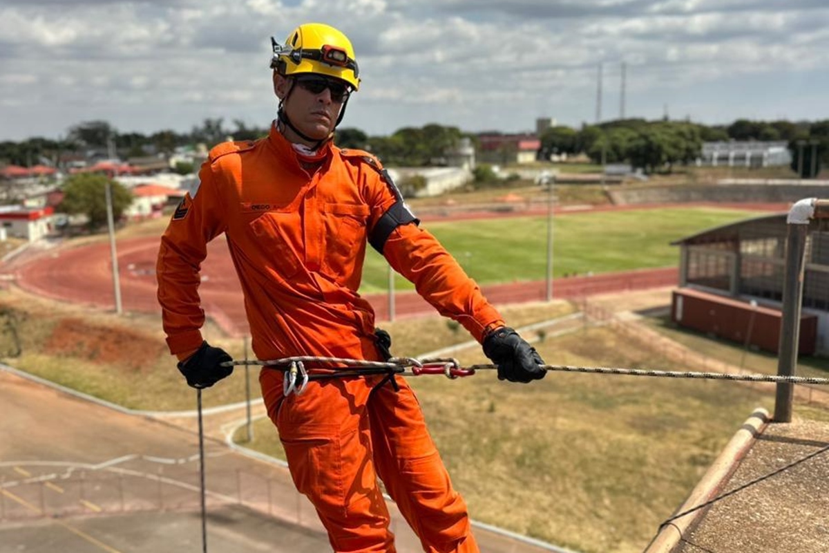 Foto colorida de homem vestido de bombeiro, pendurado por uma corda - Metrópoles.