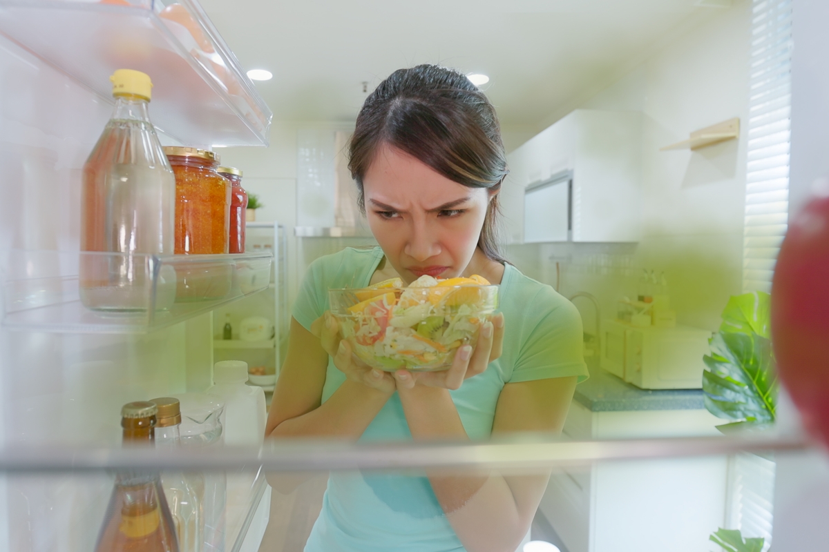 Foto colorida mulher cheirando comida que está dentro da geladeira - Metrópoles