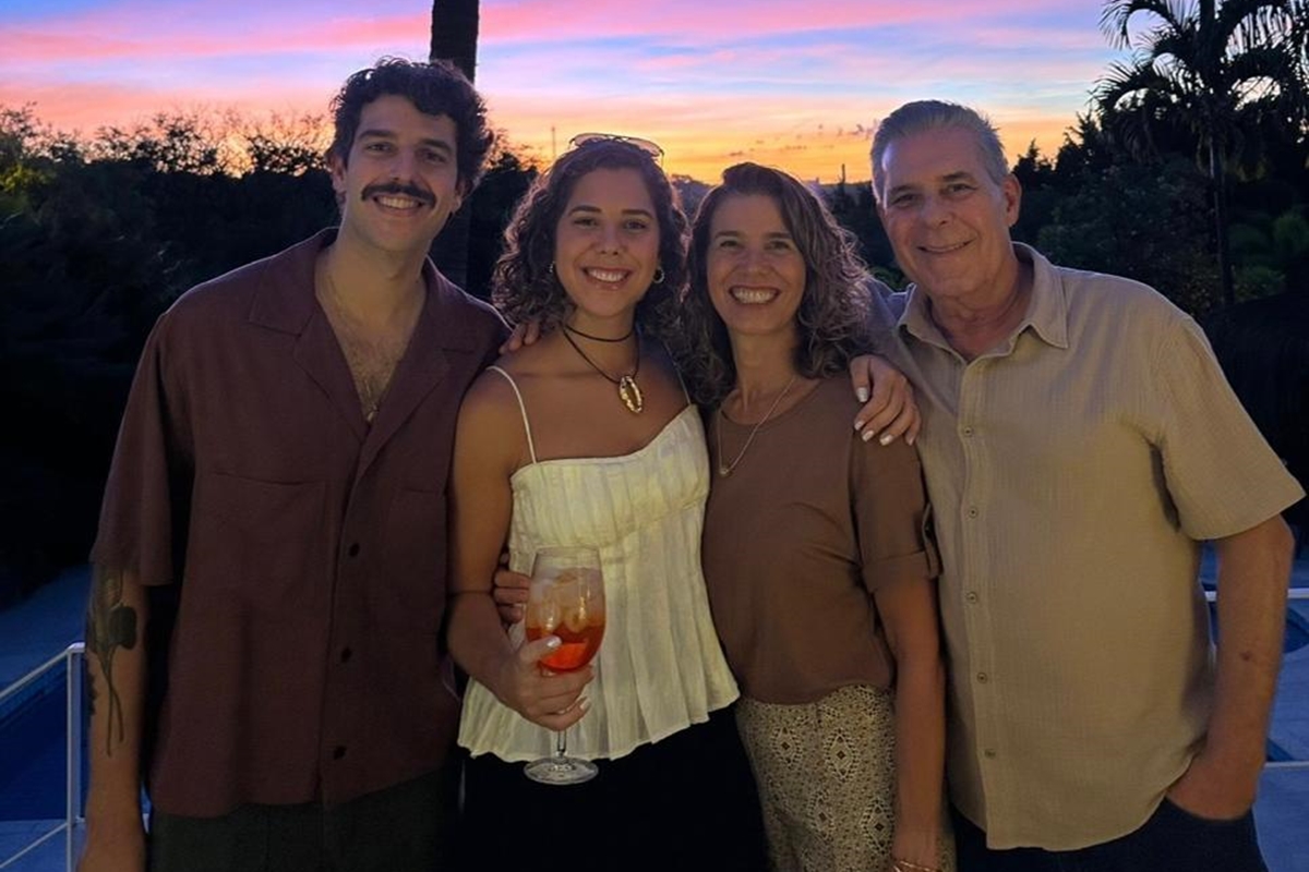 Foto colorida de família reunida se abraçando, em foto tirada em ambiente aberto, no fundo o pôr do sol - Metrópoles.